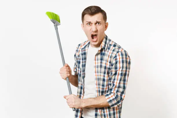 Young combative angry housekeeper man in checkered shirt holding and sweeping with green broom isolated on white background. Male doing house chores. Copy space for advertisement. Cleanliness concept