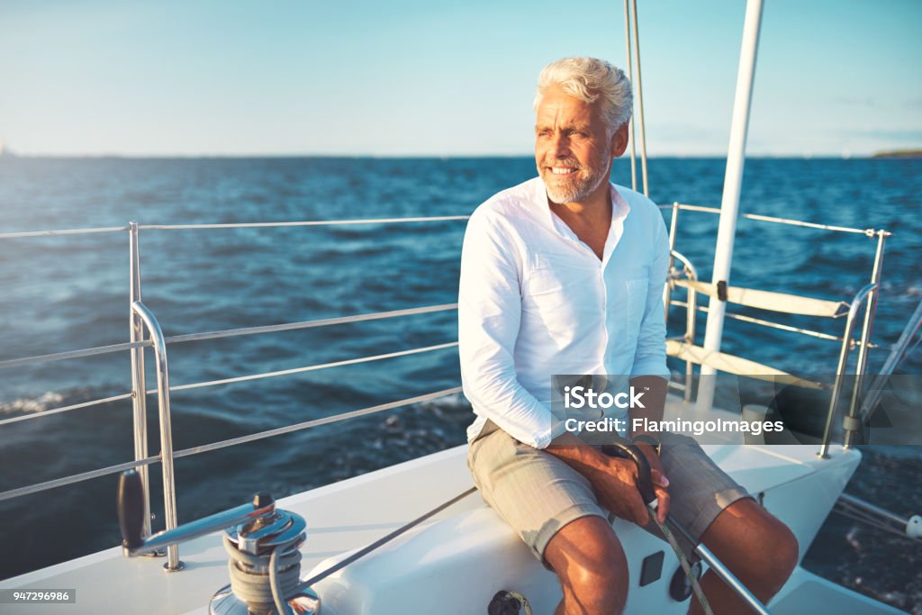 Smiling mature man sailing his yacht on a sunny day Mature man sitting on the deck of his boat enjoying a sunny day sailing on the open ocean Men Stock Photo