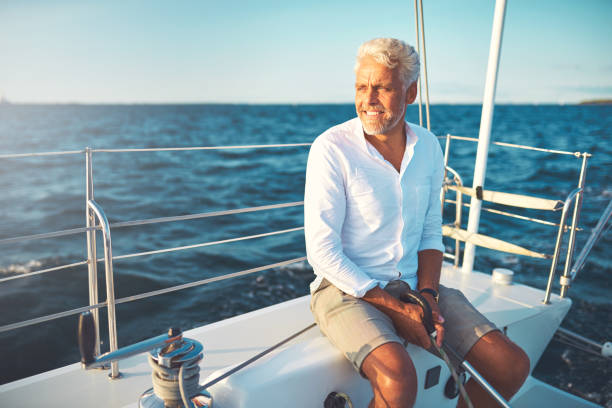 Smiling mature man sailing his yacht on a sunny day Mature man sitting on the deck of his boat enjoying a sunny day sailing on the open ocean wealthy lifestyle stock pictures, royalty-free photos & images