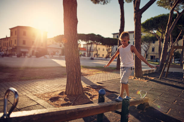 Little boy aged 7 is playing on a seesaw in the playground. The boy is walking on the see saw trying to keep balance.\nSunny summer day sunset.\nNikon D810