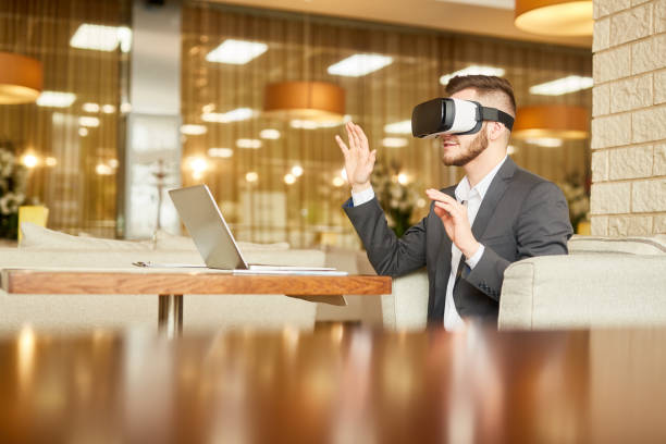 Young Caucasian manager in head-mounted display having fun while sitting at cafe table with laptop and waiting for lunch