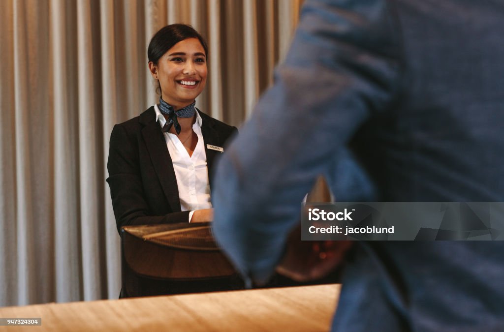 Smiling hotel receptionist attending guest at check-in counter Smiling hotel receptionist talking with male guest at reception counter. Happy female receptionist worker standing at hotel counter with businessman checking in. Hotel Stock Photo