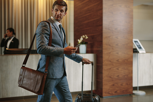 Businessman walking in hotel hallway with mobile phone and baggage. Male business traveler arriving at his hotel.