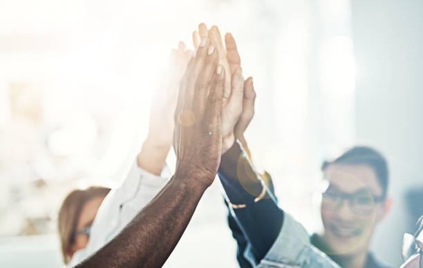 Diverse team of businesspeople high fiving together in an office Diverse team of work colleagues standing in a huddle together in a bright modern office high fiving each other dedication stock pictures, royalty-free photos & images