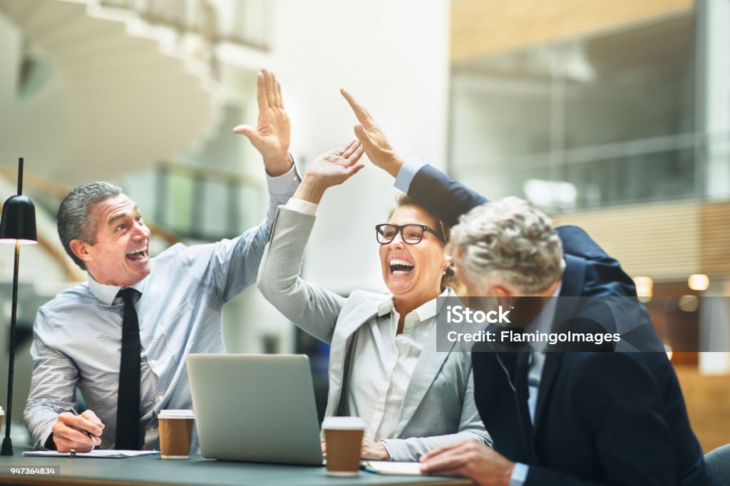 Mature businesspeople excitedly high fiving together in an office Ecstatic group of mature work colleagues high fiving together while having a meeting at a table in the lobby of a modern office building Teamwork Stock Photo