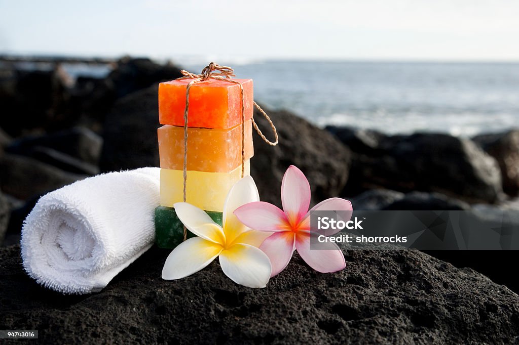 Soap and Water Stacked soaps, frangipani (plumeria, and a rolled white towel laying on a bed of lava rocks with the ocean in the background; copy space  Bar Of Soap Stock Photo