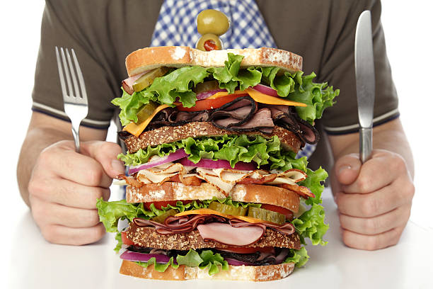 Dagwood Close up of a huge multilayered sandwich in front of a man holding a knife and fork; copy space  dagwood stock pictures, royalty-free photos & images