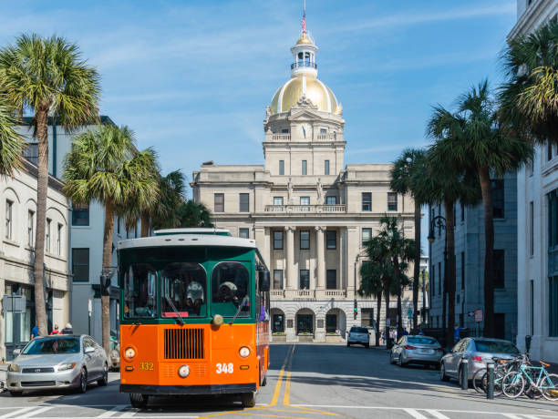 Sightseeing, Savannah, Georgia Tourists riding on one of the many orange Old Town Trolleys tour vehicles near the ornate city hall in Savannah, Georgia. trolley bus stock pictures, royalty-free photos & images