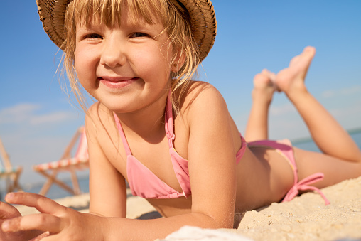 Pleased jolly cute girl in pink bikini and straw hat lying on sand and looking at camera on summer vacation