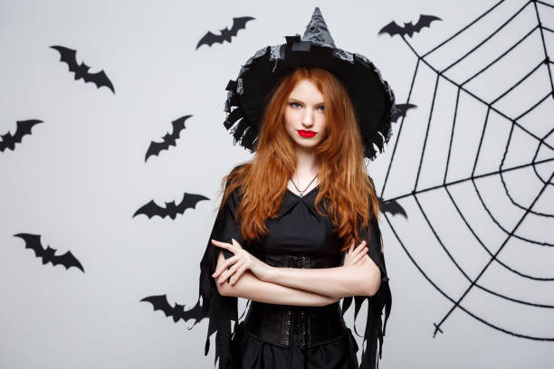 istock Halloween witch concept - Halloween Witch holding posing with serious expression over dark grey studio background with bat and spider web. 947483318