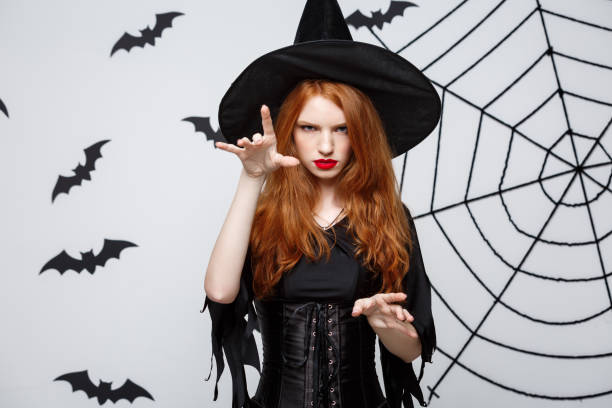 istock Halloween witch concept - Full-length Halloween Witch casting spells with serious expression over dark grey studio background with bat and spider web. 947483472