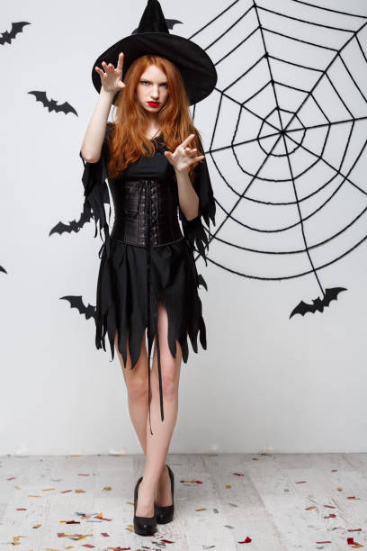 istock Halloween witch concept - Full-length Halloween Witch casting spells with serious expression over dark grey studio background with bat and spider web. 947483530