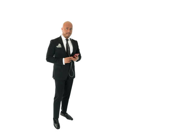 Bald businessman with black beard looking funny at camera. He is wearing an elegant black suit, a white button-down shirt and a tie. Studio shot in front of a black background.