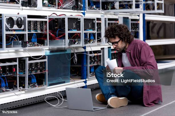 Young Computer Engineer Working While Sitting On Floor At Cryptocurrency Mining Farm Stock Photo - Download Image Now