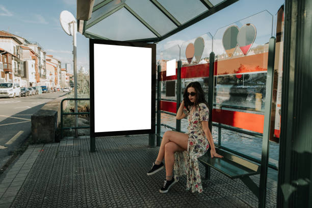 istock Young woman waiting at a bus stop with a blank billboard 947527130