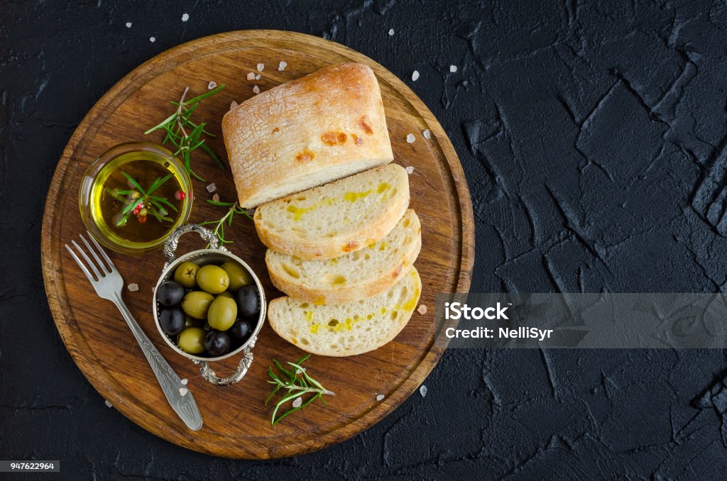 Mediterranean snacks set. Mediterranean snacks set. Green and black olives, olive oil, herbs and sliced ciabatta bread on wooden board over dark stone background. Italian food concept. Top view. Copy space. Above Stock Photo