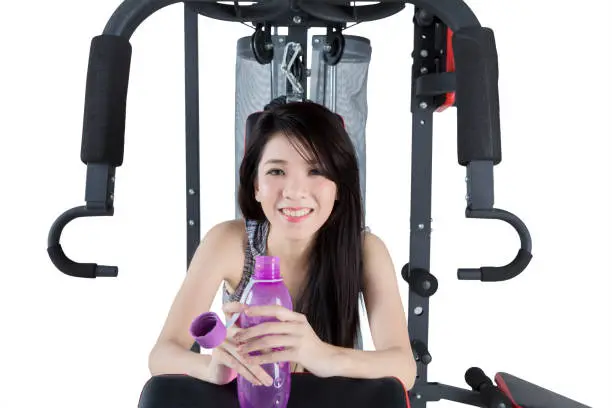Picture of happy Chinese woman holding a bottle while sitting on gym equipment, isolated on white background