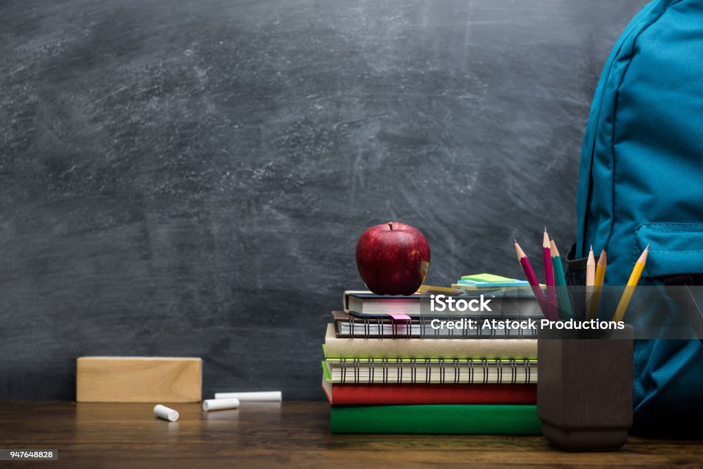 Stack of books, stationery and education supplies on wooden desk Stack of books, colorful stationery and education supplies on wooden table in classroom with blackboard in background Education Stock Photo