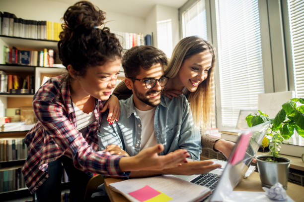 istock Group of young happy student friends is studying from a laptop while sitting at and leaning against the table near window and bookshelf in the school library. 947649346