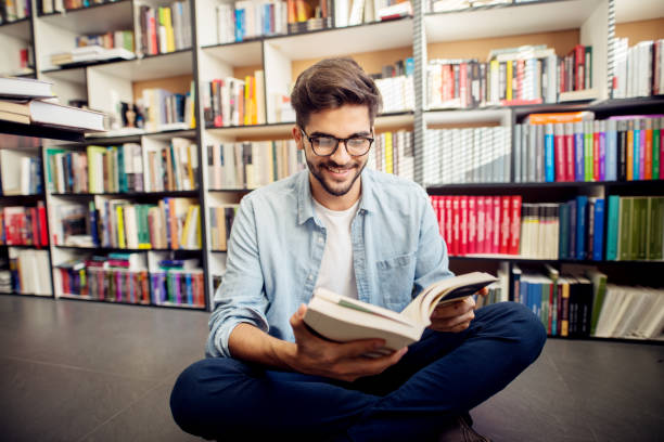 istock Front view of young hipster smiling man reading a book on the library floor. 947649418