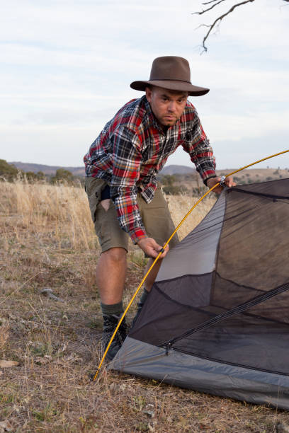 istock Camping man adjusts the side pole of his tent in a field 947655998