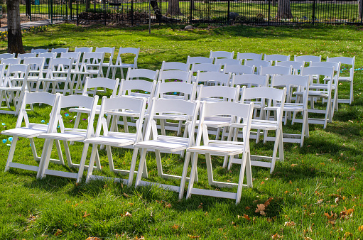 Rows of folding white chairs set up for a wedding on some green grass in a park.