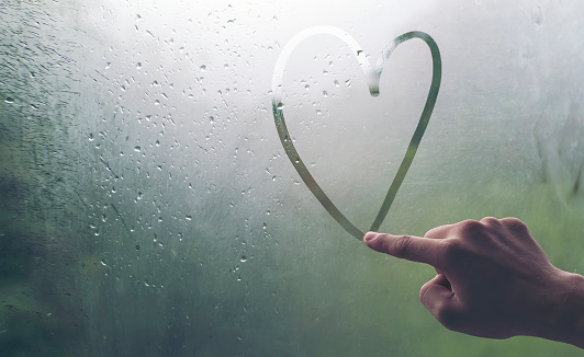 A man's hand is writing a heart-shaped glass window during a rain.