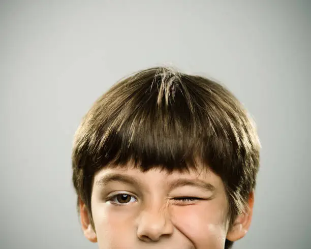 Close up half face portrait of playful caucasian real kid looking at camera. The boy is 7 years old and has brown hair. Horizontal shot of little boy winking the eye against gray background. Photography from a DSLR camera. Sharp focus on eyes.