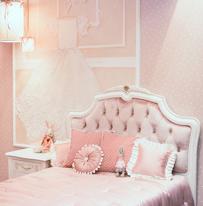 Luxury rich bedroom interior in pink color for little princess. There are different pillows on the bad, lighters on the wall and toy rabbit near the bedside table