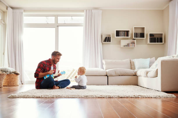 padre tocando el ukelele con hijo en su sala de estar - sentado en el suelo fotografías e imágenes de stock