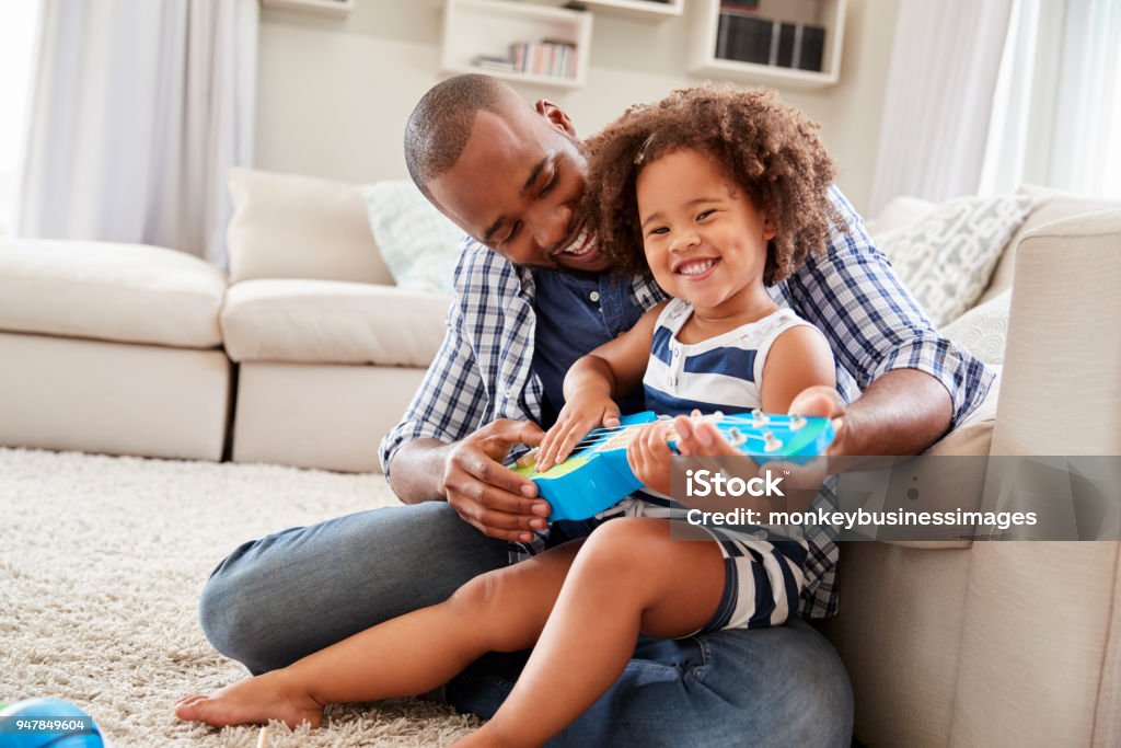 Hija de niño se encuentra en dadÕs la rodilla tocando el ukelele en casa - Foto de stock de Familia libre de derechos