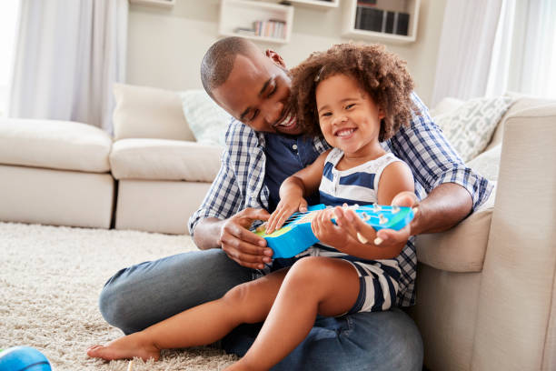 la figlia del bambino si siede sul ginocchio del papà giocando a ukulele a casa - padre single foto e immagini stock