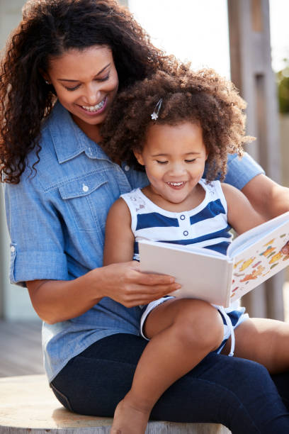 若い黒屋外 mumõs の膝の上に座って本を読んでいる女の子 - child reading mother book ストックフォトと画像