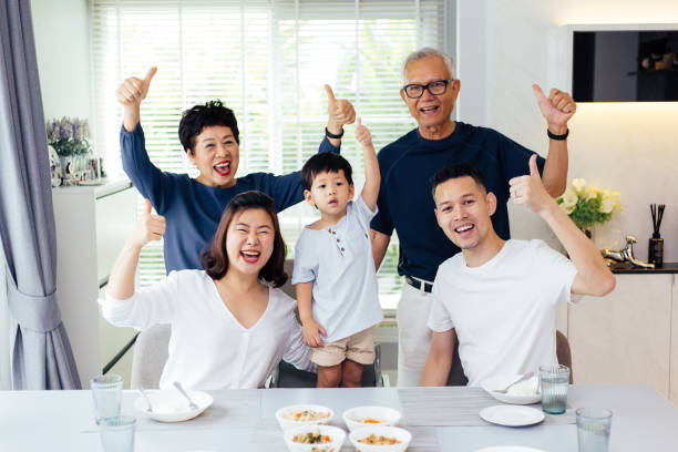 asiatische großfamilie aus drei generationen, die eine mahlzeit zusammen und zeigt daumen sich zu hause mit glück - daumen hoch fotos stock-fotos und bilder
