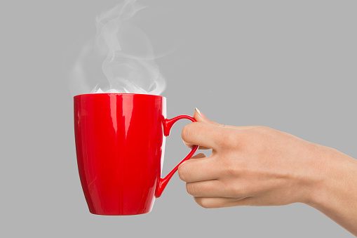 Female hand holding red cup with hot coffee at gray background, copy space