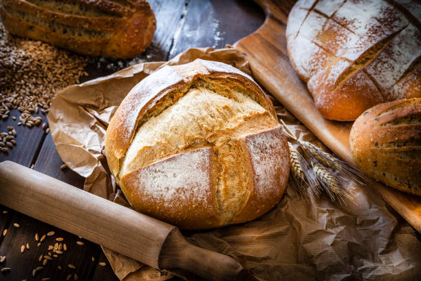 Loaf of bread still life Different types of bread shot on rustic wooden table. A fresh baked loaf of bread is on foreground on a brown craft paper. Predominant color is brown. Low key DSRL studio photo taken with Canon EOS 5D Mk II and Canon EF 100mm f/2.8L Macro IS USM baked stock pictures, royalty-free photos & images