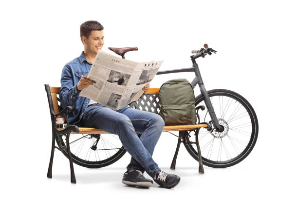 istock Young guy with a bicycle and a backpack sitting on a wooden bench and reading a newspaper 947887298