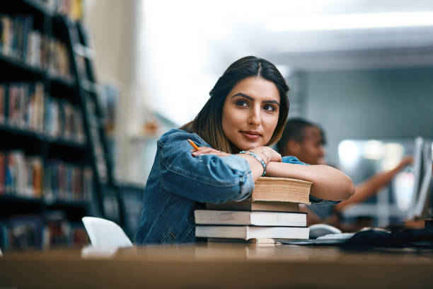 Where dreams are turned into reality Shot of a young woman resting on a pile of books in a college library and looking thoughtful day dreaming stock pictures, royalty-free photos & images