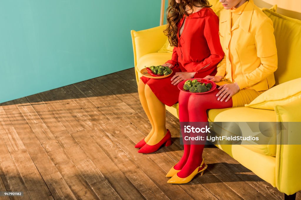 cropped image of retro styled girls in colorful dresses sitting with plates of broccoli at home Living Room Stock Photo
