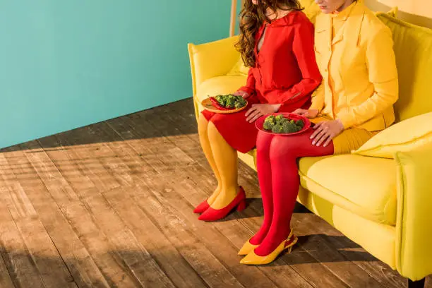 Photo of cropped image of retro styled girls in colorful dresses sitting with plates of broccoli at home