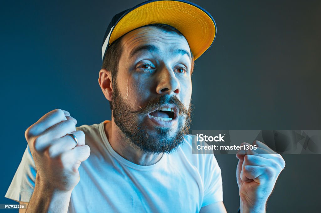 Sports, emotions and fan people concept - sad man watching sports on tv and supporting team at home Sports, fan human emotions and people concept - sad young man watching sports on tv and supporting team at home. Crying emotional man screaming in colorful bright lights at studio. Facial expression concept. Fan - Enthusiast Stock Photo