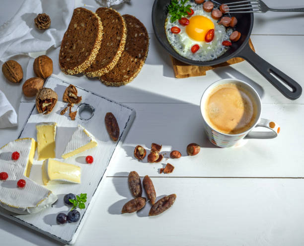 round Camembert cheese on a white board, fried eggs with sausage and a cup of coffee on a white wooden table, top view