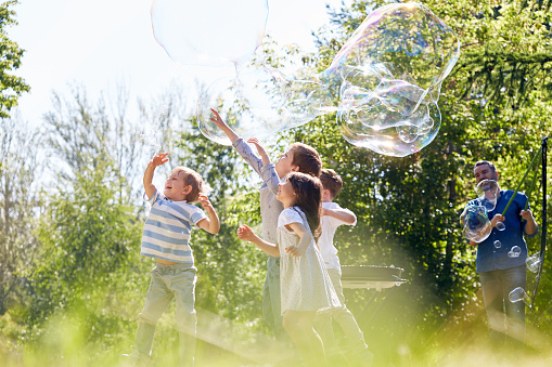 Profile view of little friends having fun while participating in soap bubble show at green public park illuminated with sunbeams