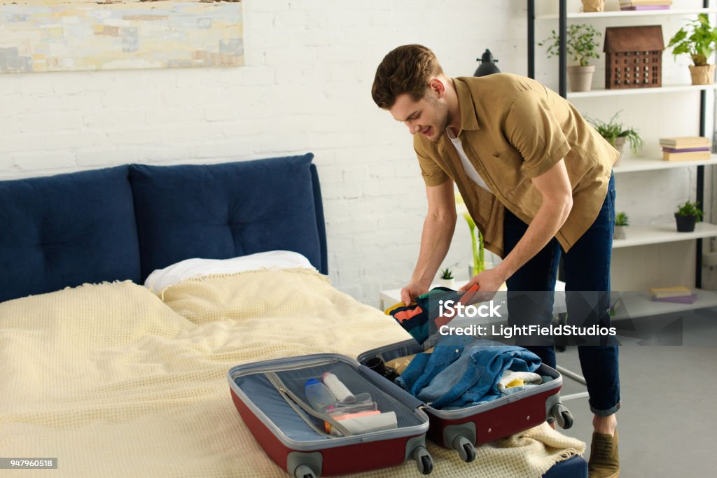 young smiling man packing clothes into travel bag Packing Stock Photo