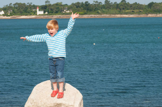 Happy 5 year old stands on rock waving hands with sea behind stock photo
