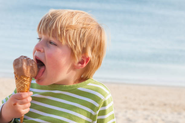 glücklich 5 jahre alten jungen blonden jungen lecken große schokoladeneis in einem kegel mit sandstrand und blaues meer hinter ihm - child chocolate ice cream human mouth stock-fotos und bilder