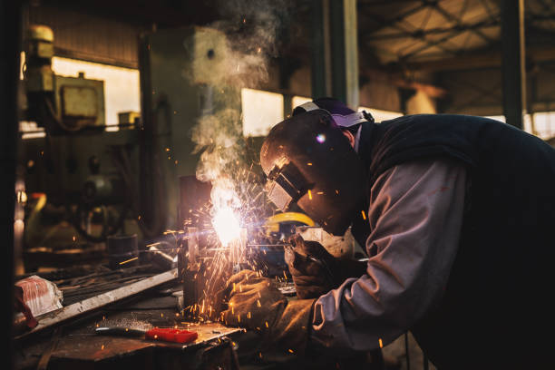 istock Welder in protective uniform and mask welding metal pipe on the industrial table while sparks flying. 947972058