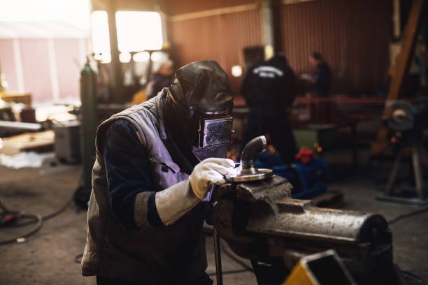 istock Welder in protective uniform and mask welding metal pipe on the industrial table while sparks flying. 947977168