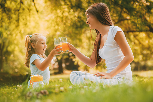 Beautiful Mother and daughter at the park having little picnic. They are enjoying and drinking orange juice.
