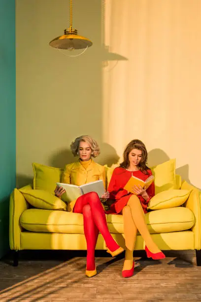 Photo of women in retro clothing reading books sitting on yellow sofa at bright apartment, doll house concept